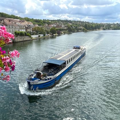 Blauw-wit riviercruiseschip MS Fleur varend op kalme rivier in Noord-Frankrijk, met roze bloemen op voorgrond en huizen aan heuvels.
