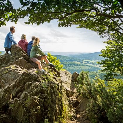 Vier wandelaars zitten op een grote rotsformatie langs de Rennsteig, uitkijkend over beboste heuvels en valleien onder bewolkte hemel.