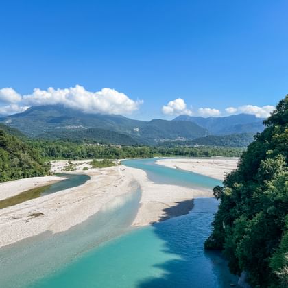Tagliamento-bedding met turquoise waterkanalen door witte grindbanken, omgeven door groene bossen en bergen onder blauwe lucht.