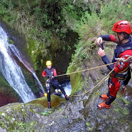 Twee personen met rode helmen en wetsuits abseilen langs rotswanden naast een waterval op Madeira tijdens canyoning.