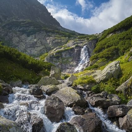 Bergstroom stroomt over rotsen in het Tatra-gebergte met waterval die groene hellingen afdaalt onder blauwe lucht met witte wolken.