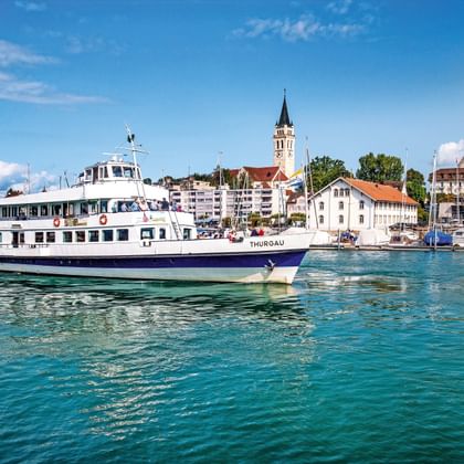 Witte passagiersveerboot Thurgau varend op turquoise water van Bodenmeer met haven van Romanshorn, kerktoren en jachthaven op achtergrond.