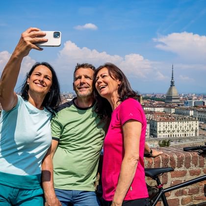 Drie fietsers maken een selfie op een bakstenen muur met uitzicht op Turijn, Italië. De Mole Antonelliana toren is zichtbaar in de stadshorizon.
