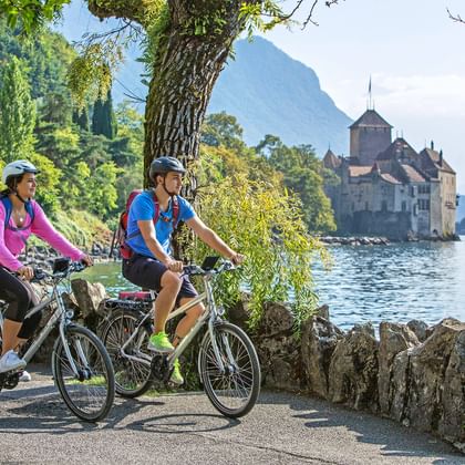 Twee fietsers op meerpad met kasteel Chillon aan Meer van Genève. Vrouw in roze shirt en man in blauw shirt met fietsen, bergen op achtergrond.