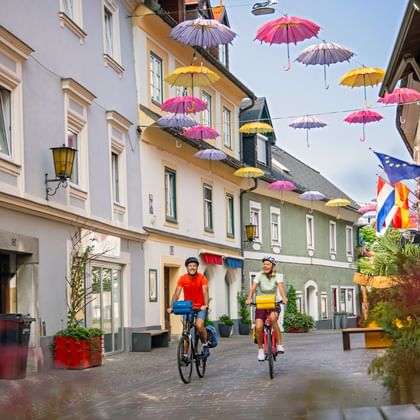 Twee fietsers rijden door een voetgangersstraat in Villach, Karinthië. Kleurrijke paraplu's hangen boven tussen historische gebouwen.