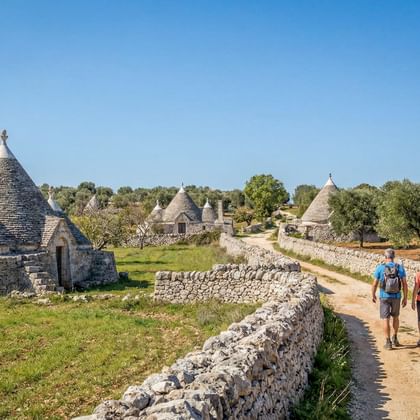 Twee wandelaars op een zandpad tussen stenen muren naar traditionele trulli-huizen met kegelvormige daken in Apulië onder blauwe hemel.