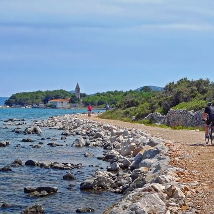 Fietsers rijden op een grindpad langs de kust van Losinj met rotsachtige kustlijn, heldere blauwe zee en historische kerktoren in de verte.