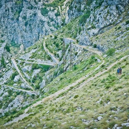 Twee wandelaars op een grasrijk bergpad met uitzicht op dramatische serpentines in rotsachtige kliffen in de Baai van Kotor, Montenegro.