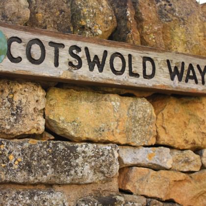 Houten Cotswold Way bewegwijzering met pijl op verweerde honingkleurige stenen muur. Het bord heeft zwarte letters en een rond logo.