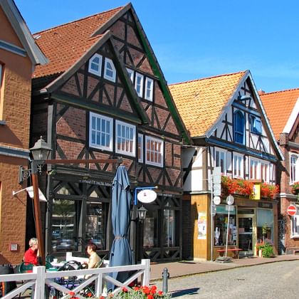 Rij kleurrijke vakwerkhuizen met rode pannendaken op de fietsroute Magdeburg-Hamburg. Mensen zitten aan café-tafels buiten.