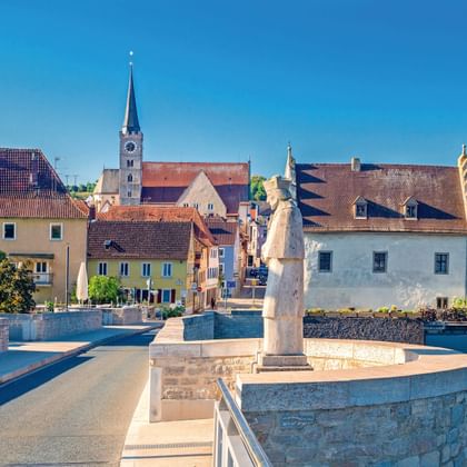 Stenen brug met standbeeld in Ochsenfurt aan de Main. Historisch stadscentrum met kerktoren en kleurrijke gebouwen onder blauwe lucht.