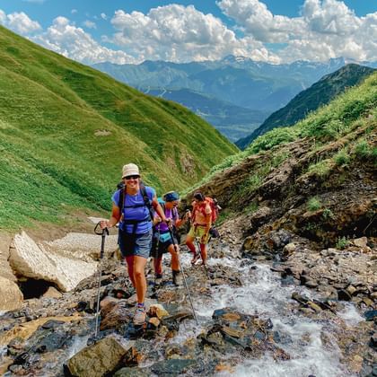 Groep wandelaars met rugzakken en wandelstokken steekt rotsachtige beekbedding over in Georgische bergen met groene heuvels.