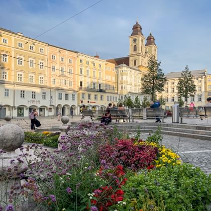Linz Hauptplatz met kleurrijke bloembedden op de voorgrond, gele historische gebouwen en kerk met twee torens onder blauwe lucht.