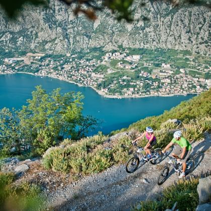 Twee mountainbikers op een grindpad met uitzicht op de baai van Kotor in Montenegro, met blauw water, kustplaats en bergen.