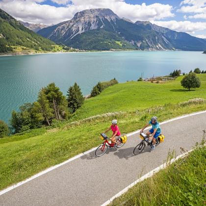 Twee fietsers op een weg naast het Reschenmeer met turquoise water, groene weiden en dramatische Alpenbergen onder een bewolkte hemel.