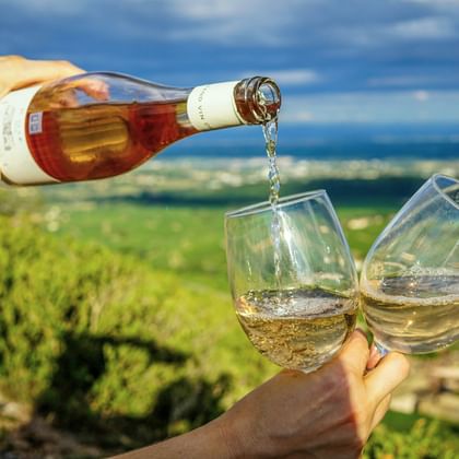 Hand schenkt witte wijn uit een fles in twee glazen met wijngaarden en blauwe lucht op de achtergrond in de Provence, Zuid-Frankrijk.