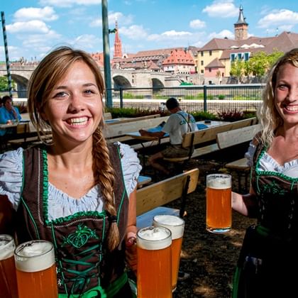Twee glimlachende vrouwen in traditionele Duitse dirndl houden meerdere bierglazen vast in een biertuin met historische gebouwen op de achtergrond.