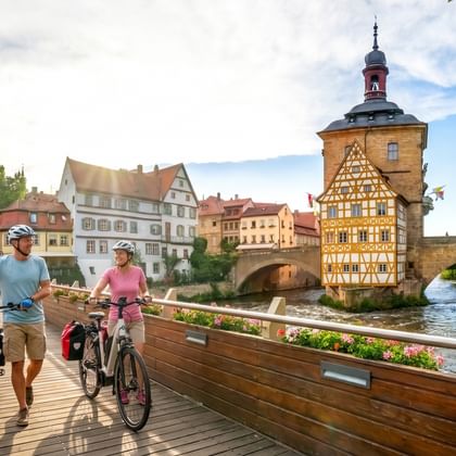 Twee fietsers lopen met hun fietsen over een houten brug in Bamberg, met het historische vakwerken stadhuis aan de rivier op de achtergrond.