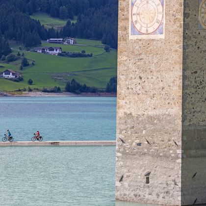 Beroemde kerktoren uit Reschenmeer met zichtbare klokken. Drie fietsers rijden langs het pad aan het meer met bergen op de achtergrond.