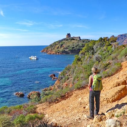 Wandelaar met groene rugzak op rotsachtig kustpad met uitzicht op turquoise Middellandse Zee bij Girolata, Corsica, historisch fort zichtbaar.