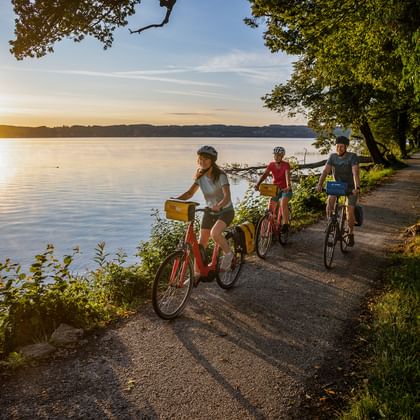 Drie fietsers met helmen op oeverpad aan het Starnberger See tijdens gouden zonsopgang, omringd door herfstbomen en kalm water.