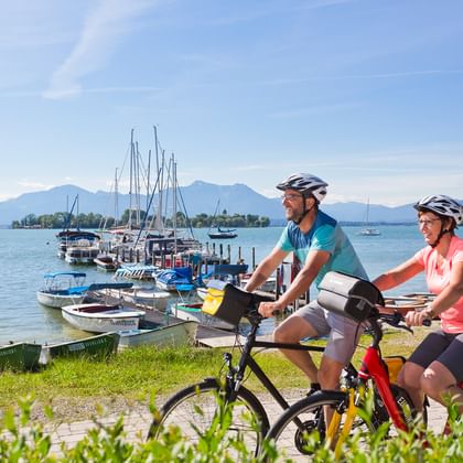 Twee fietsers met helmen pauzeren bij het Chiemsee bij de jachthaven van Gstadt. Bergen en zeilboten zijn zichtbaar over het blauwe water.