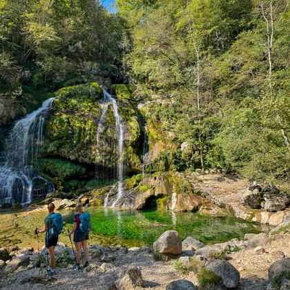 Twee wandelaars met rugzakken en wandelstokken bij een helder groen bassin bij de Virje-waterval, omringd door bemoste rotsen en bos.