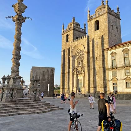 Porto Cathedral's twin towers and baroque facade with ornate stone pillar in foreground. Tourists and cyclists gather in the square.