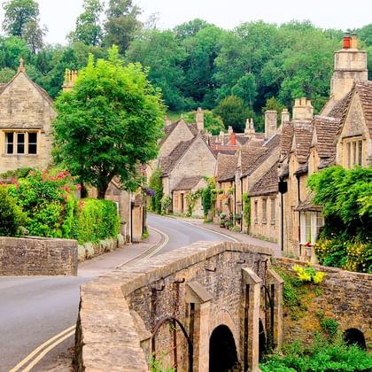 Historisch Cotswold dorp met honingkleurige stenen huizen en middeleeuwse brug. Weg kronkelt door dorp met groene bomen en tuinen.