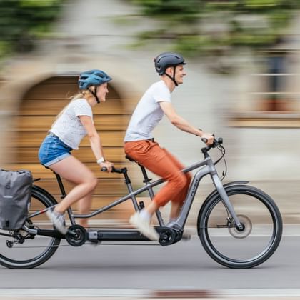 Twee fietsers op een grijze tandem e-bike met bewegingsonscherpte achtergrond. Vrouw met blauwe helm en spijkershort, man met donkere helm.