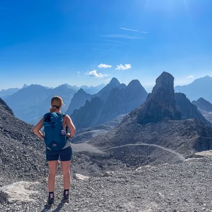 Wandelaar met blauwe rugzak op rotsachtig bergpad in de Allgäuer Alpen, kijkend naar dramatische bergtoppen onder heldere hemel.
