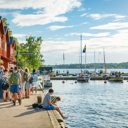 Haven van Fjäderholmarna met bezoekers op de waterkantpromenade. Rode houten gebouwen langs de kust, boten in het water, Zweedse vlaggen zichtbaar.