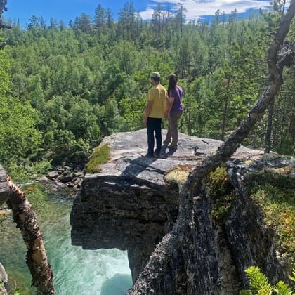Twee personen in gele en paarse kleding staan op rotsklif met uitzicht op dicht groen bosdal in Dovrefjell, Noorwegen.