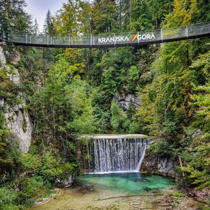 Hangbrug met opschrift Kranjska Gora over een beboste kloof boven een waterval die uitmondt in een turquoise poel omgeven door groen.