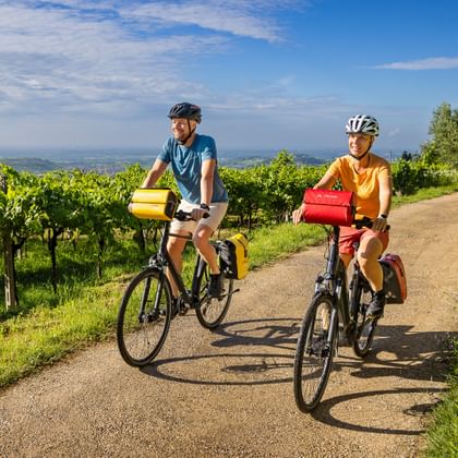 Twee fietsers met fietstassen rijden over grindpad door groene wijngaarden bij Gardameer, met blauwe lucht en verre heuvels op achtergrond.