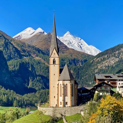 Historische kerk met hoge toren in Heiligenblut am Großglockner, omgeven door traditionele Alpenhuizen en besneeuwde bergen.