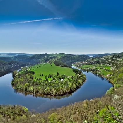 Luchtfoto van de Moldau die een dramatische hoefijzerbocht vormt door beboste heuvels en groene weiden onder een blauwe hemel met wolken.