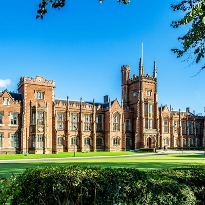 Rood bakstenen neogotisch gebouw van Queen's University Belfast met torens en spitsen, omgeven door verzorgde gazons en heggen.