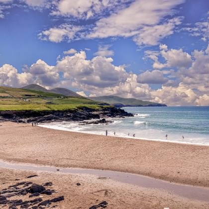 Zandstrand langs de Kerry Way in Ierland met wandelende mensen, rotsformaties, groene heuvels en dramatische bewolkte lucht boven blauwe oceaan.