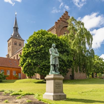 Bronzen standbeeld van Hans Christian Andersen op wit voetstuk in park met kerktoren en historische bakstenen gebouwen in Odense, Denemarken.
