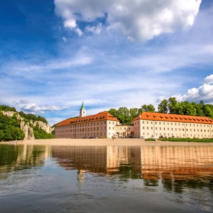 Klooster Weltenburg met rode pannendaken en groene toren weerspiegeld in de Donau, omgeven door kalksteenrotsen en bos.
