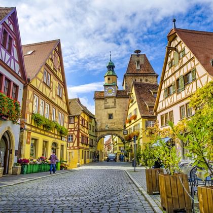 Geplaveide straat in oude stad Rothenburg met kleurrijke vakwerkhuizen en middeleeuwse poorttoren met klok onder blauwe lucht.