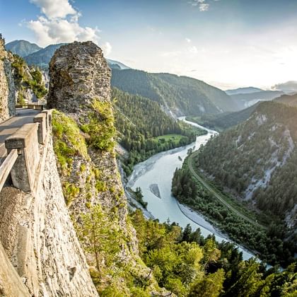 Kronkelende bergweg uitgehouwen in rotswand boven de Rijnkloof in Graubünden. Rivier stroomt door bebost dal met rotsformaties.