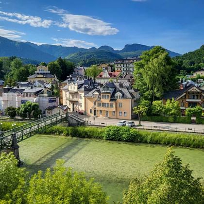 Uitzicht op Bad Ischl met een groene voetgangersbrug over een rivier, kleurrijke gebouwen en bergketens onder een blauwe lucht met wolken.