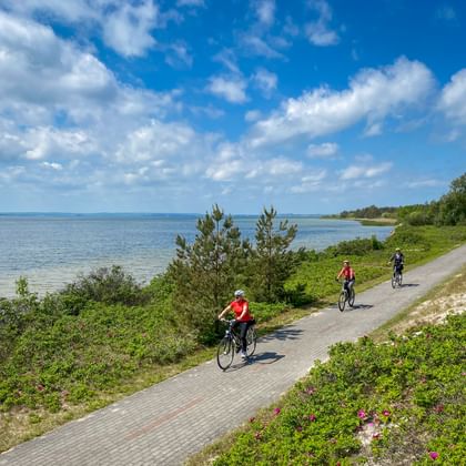 Drie fietsers op een verhard kustpad langs de Oostzee tussen Stettin en Danzig, met blauw water, groene vegetatie en wolken.