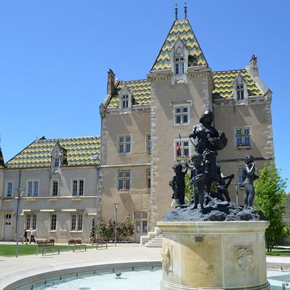 Stenen kasteel in Côtes de Beaune met kenmerkende groen-gele dakpannen, toren en sierlijke architectuur met bronzen standbeeld op de voorgrond.