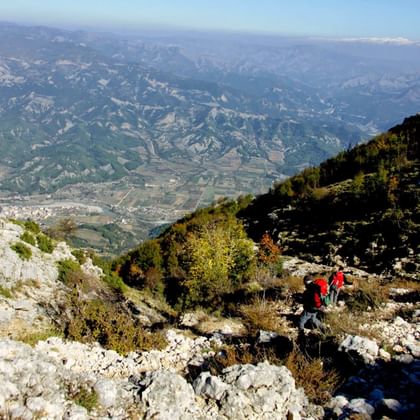 Twee wandelaars met rode jassen op rotsachtig bergpad in Hidden Valley, Albanië, met uitgestrekt uitzicht op vallei en verre bergen.