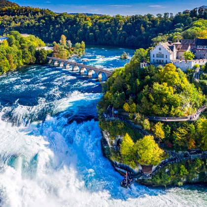 Luchtfoto van de Rijnwaterval bij Schaffhausen met waterval, historische gebouwen, stenen brug en groene bossen onder blauwe hemel.