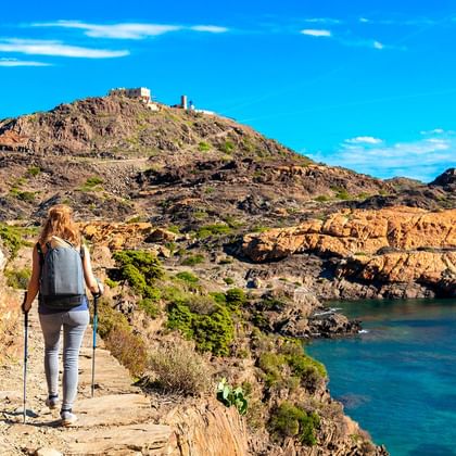 Vrouwelijke wandelaar met rugzak en wandelstokken op rotsachtig kustpad met uitzicht op turquoise Middellandse Zeebaai met ruige kliffen.