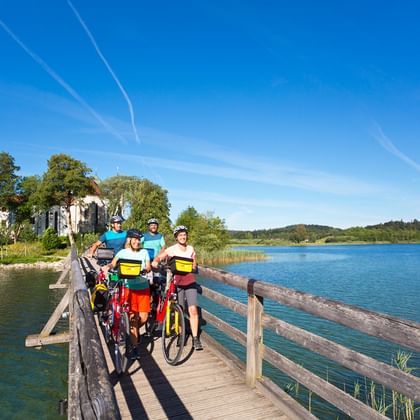Vier fietsers met fietsen op een houten brug over een meer bij klooster Seeon. Blauwe lucht en groene heuvels op de achtergrond.